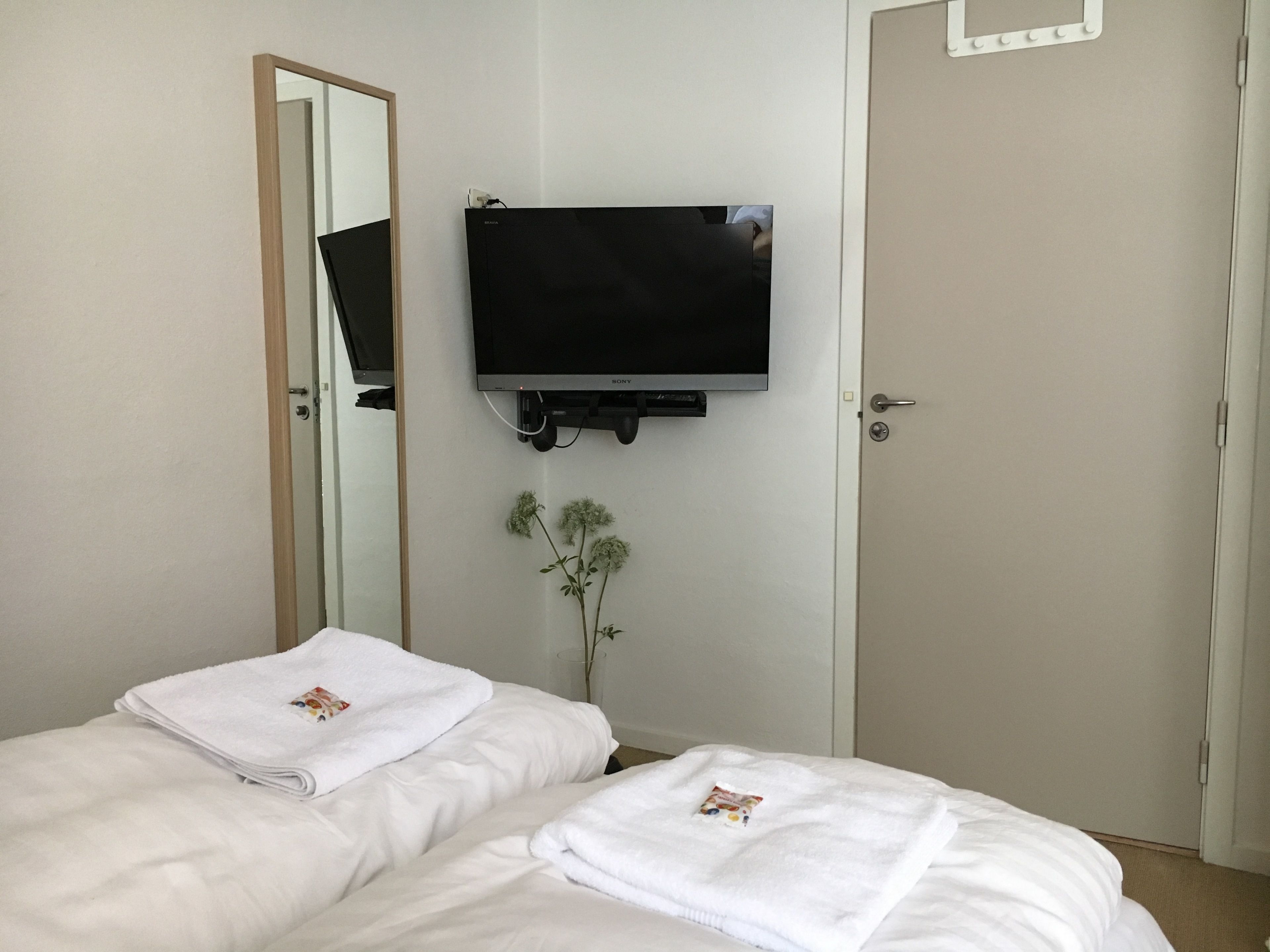 Ein Hotelzimmer mit zwei Betten, einem an der Wand befestigten Fernseher, einem Spiegel und einer Tür.