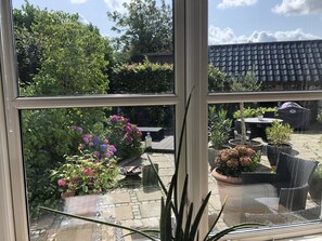 A view through a window of a patio area with potted plants, outdoor furniture, and a tiled roof in the background. - Helts B&B / Helts Guesthouse (Herning)