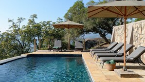 A poolside area with lounge chairs and umbrellas, a stone wall, and a view of the landscape. - Tomjachu Bush Retreat (Mbombela)