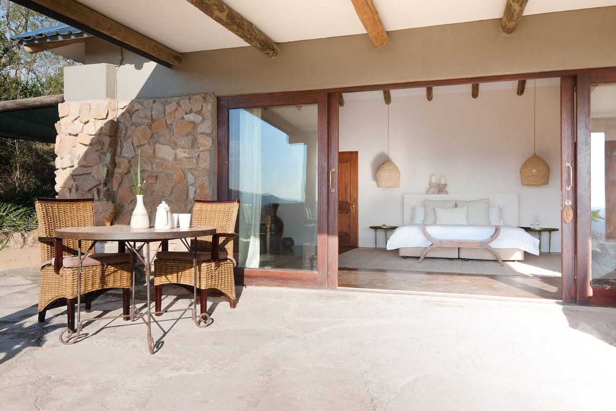 An outdoor patio with wicker furniture, a stone wall, and a view of a bedroom with a bed and bedside lamps.