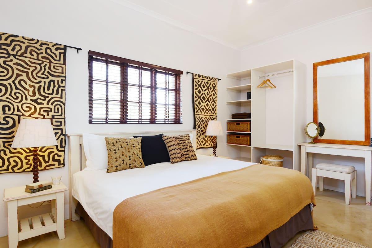 A well-lit bedroom with a large bed, a wardrobe, a vanity with a mirror, and a window with blinds.