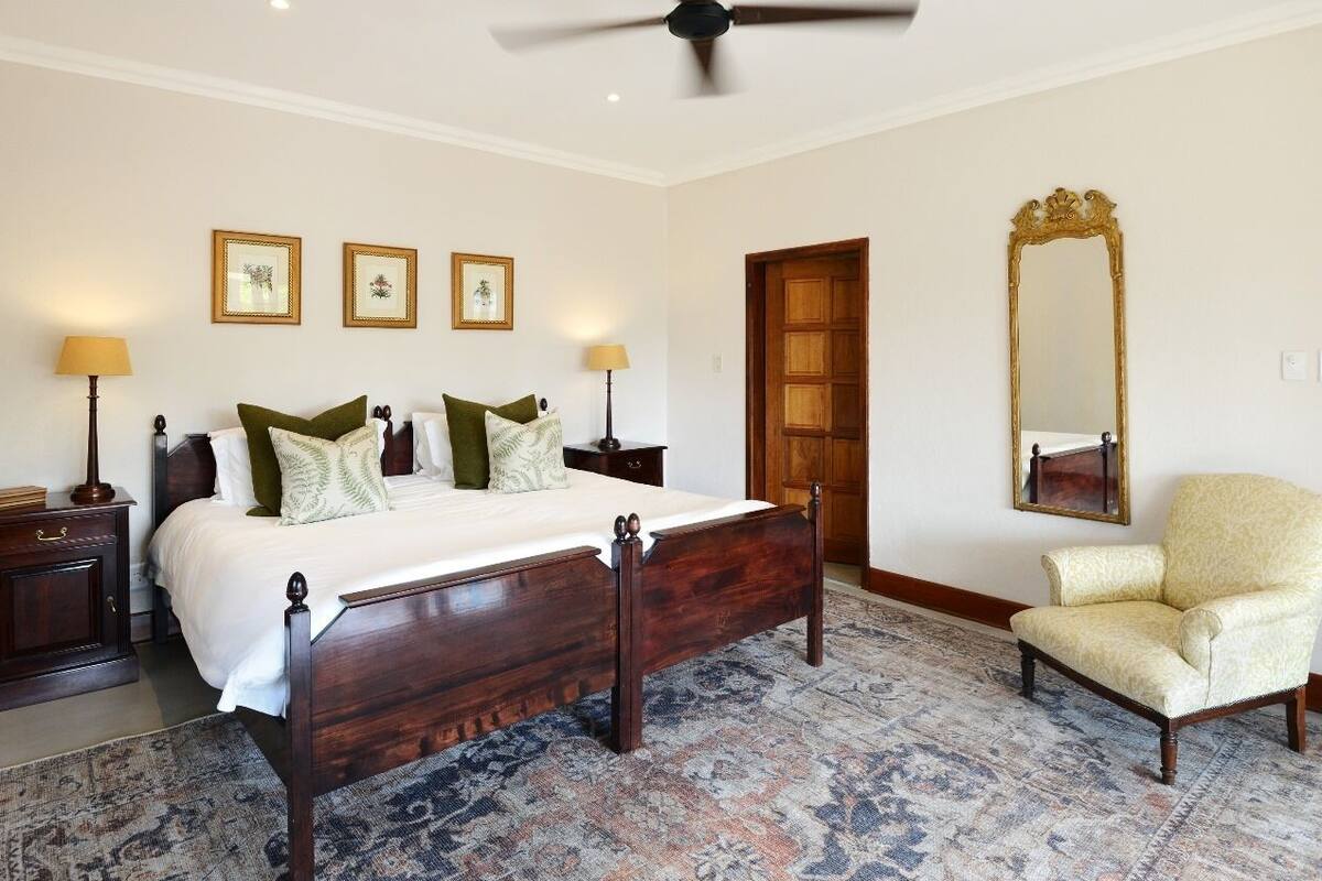 A bedroom with a wooden bed, two bedside tables with lamps, a chair, a large mirror, and three framed pictures on the wall.