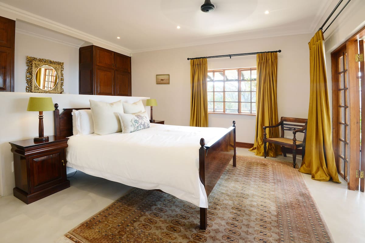 A bedroom with a large bed, wooden headboard, a nightstand, a lamp, a chair, a window with curtains, and a patterned rug.