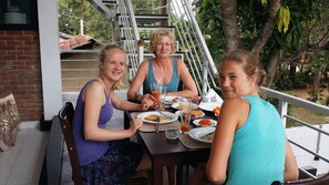 Three people dining at an outdoor table with plates of food, glasses, and a metal railing in the background.