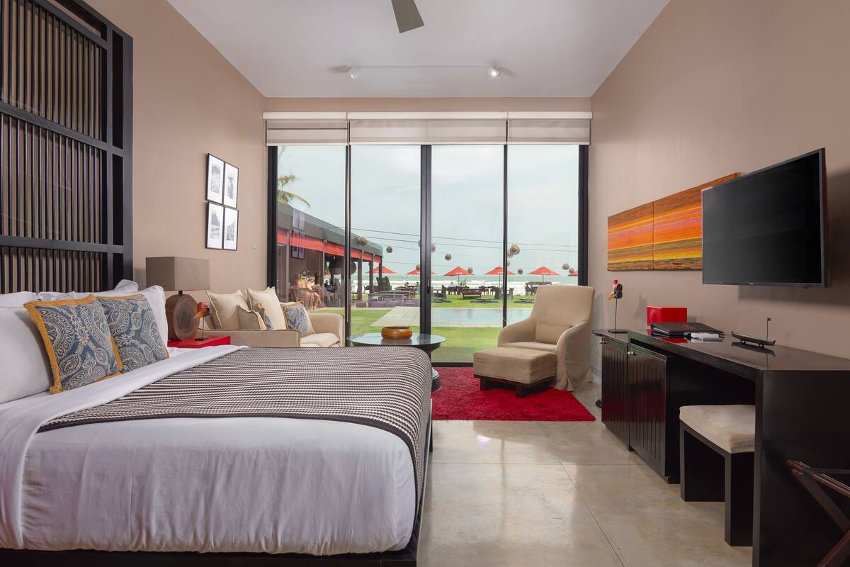 A modern hotel room with a large bed, a desk, a TV, and a balcony with a view of the pool and outdoor area.