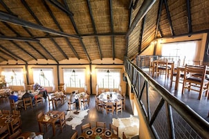 Uma sala de jantar ampla com pé direito alto e grandes janelas, com várias mesas e cadeiras.