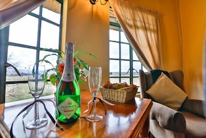 Uma mesa com uma garrafa de vinho J.C. Penney, dois copos, uma cesta de frutas e vista do exterior através de grandes janelas.