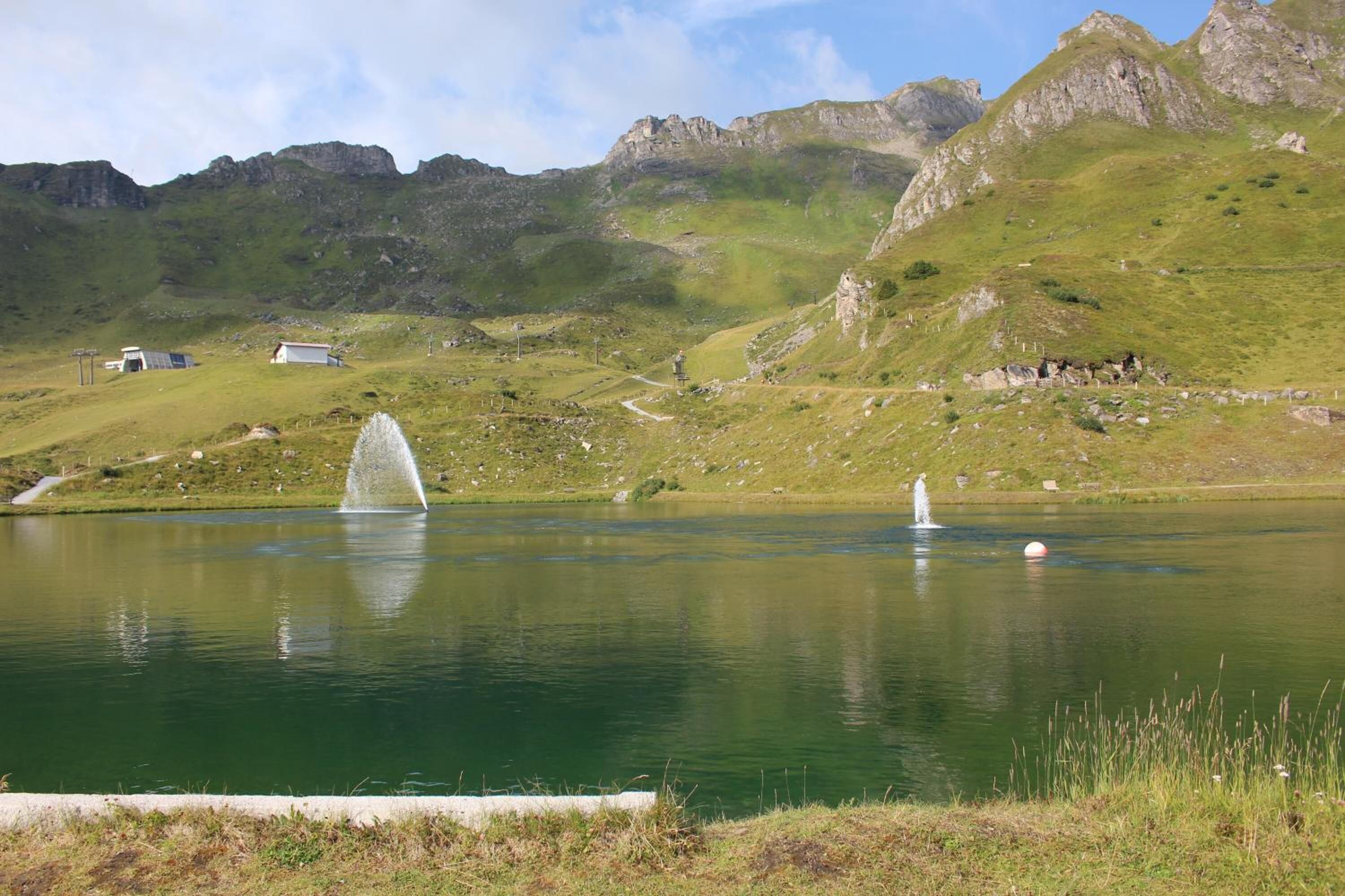 Ein ruhiger See mit einer Springbrunnen, umgeben von grünen Hügeln und einigen Gebäuden im Hintergrund.