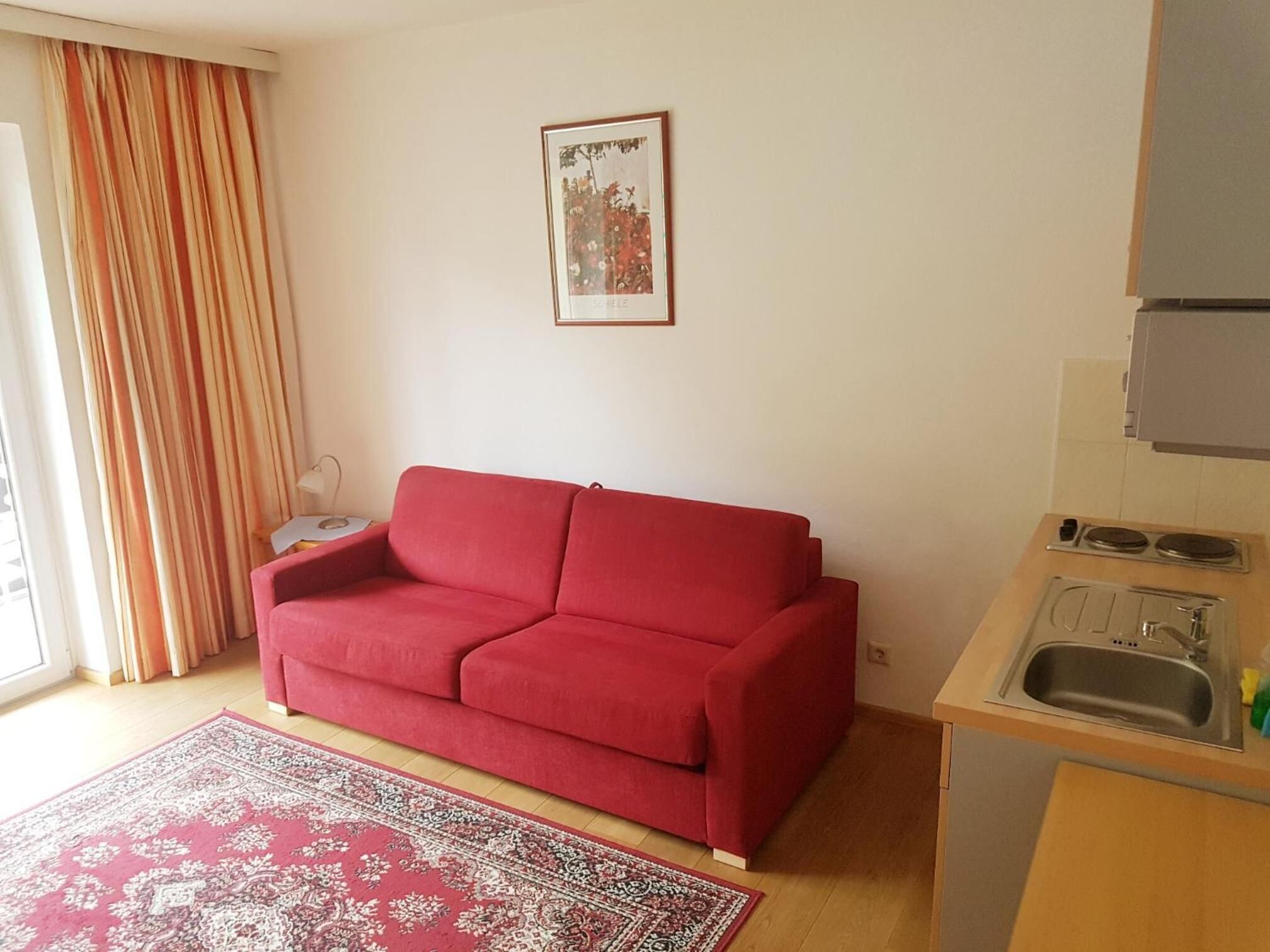 A compact living space with a red sofa, a kitchen area with a sink and stove, a patterned rug, and a window with curtains.
