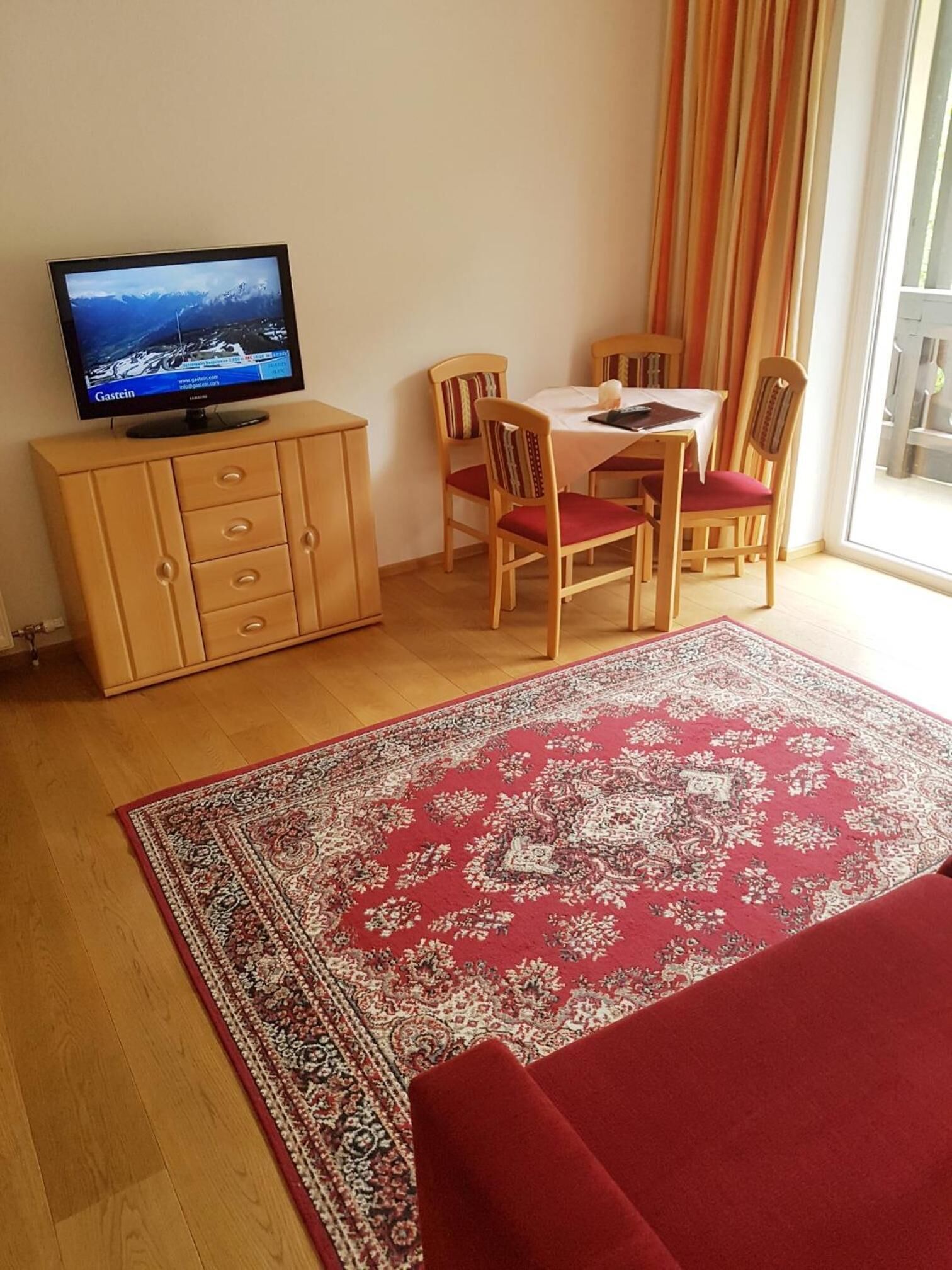 A room with a television on a wooden cabinet, a dining table with chairs, and a large patterned rug.