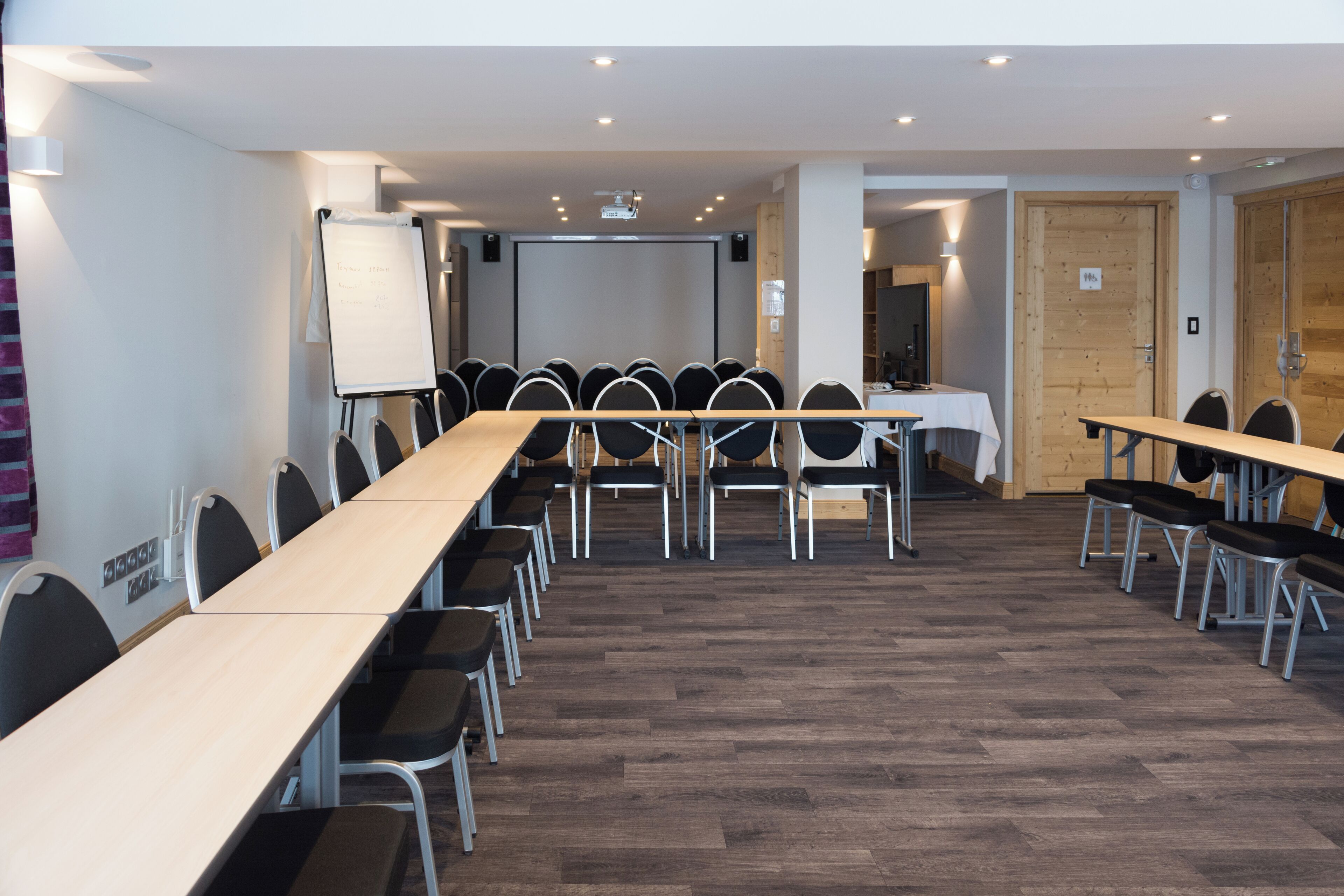 A conference room with a long table, multiple chairs, a whiteboard, and a projection screen.