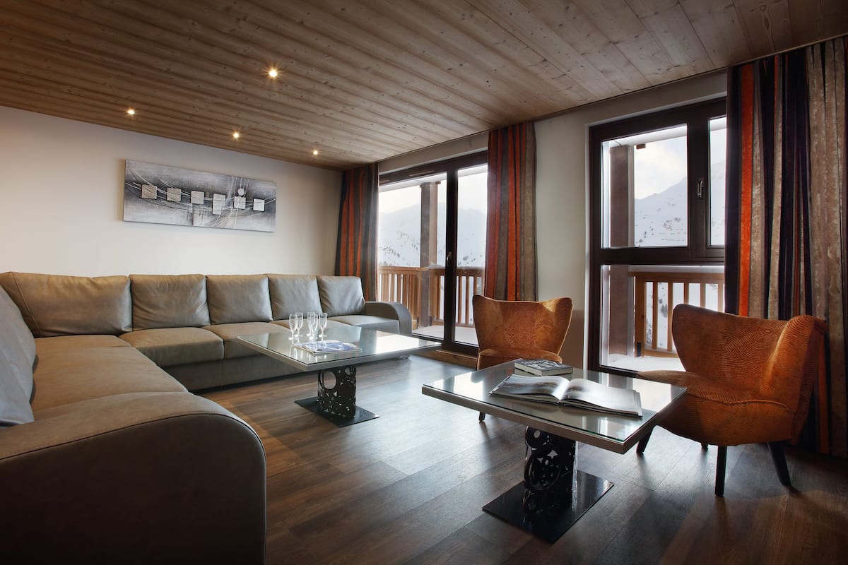 Una sala de estar moderna con un sofá modular, una mesa de centro de vidrio y dos sillones. Se aprecia una ventana grande con vistas a un paisaje nevado.
