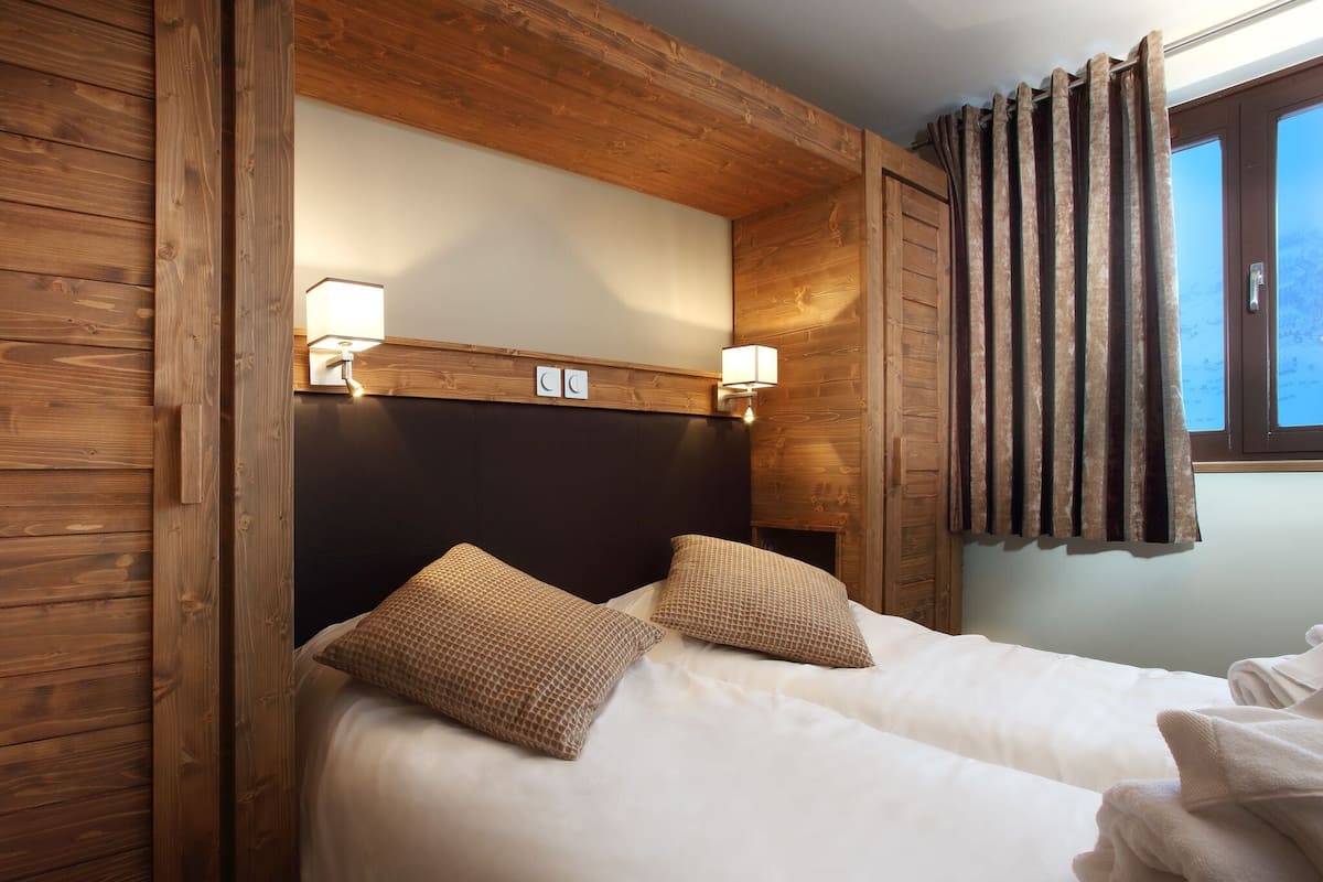 Cama doble con dos almohadas, paneles de madera en las paredes, cabecera con dos luces fijadas a la pared y una ventana con cortinas.