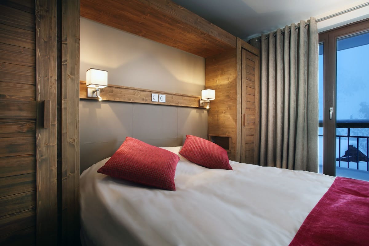 Habitación de hotel con cabecera de madera, dos lámparas de noche, cama con ropa blanca y almohadas rojas, y vistas al exterior a través de una puerta corrediza de vidrio.