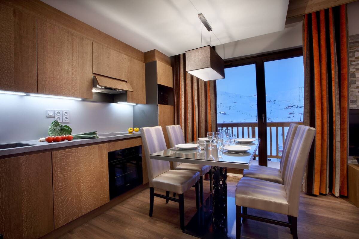 Una cocina moderna con armarios de madera, un comedor con mesa de vidrio y vistas a un paisaje nevado a través de amplias ventanas.