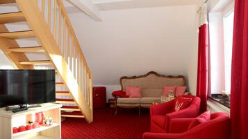A room with a wooden staircase, a sofa set, a red armchair, a television on a stand, and a window with red curtains.