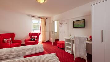 A room with a red carpet, a red armchair with a green pillow, a white desk with a wicker chair, and a wardrobe.