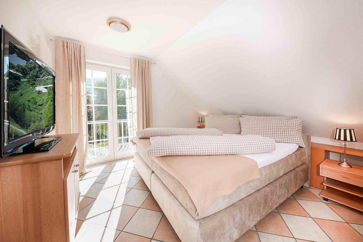 A bedroom with a bed, a television on a wooden cabinet, a nightstand with a lamp, and a window with curtains.