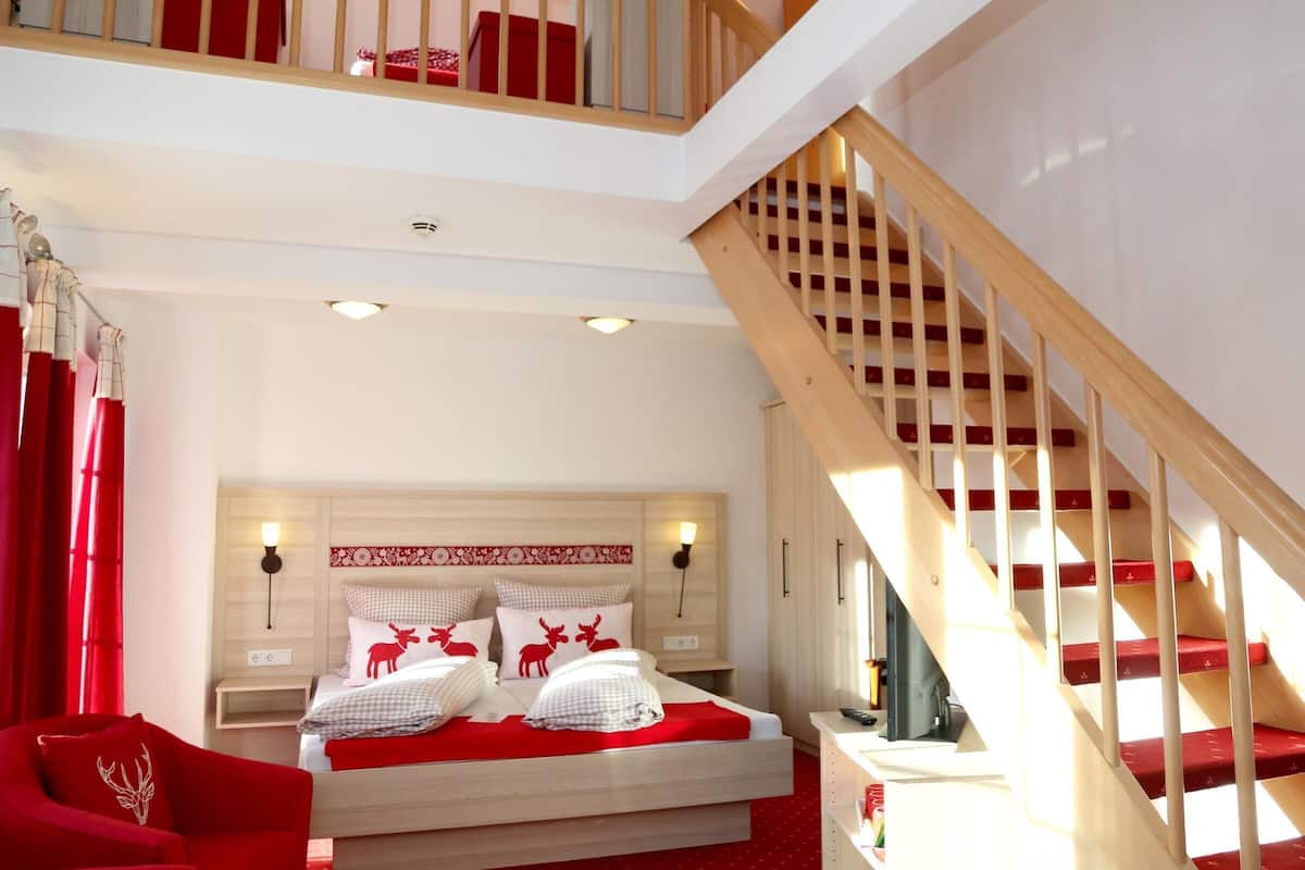 A modern hotel room with a wooden staircase, a large bed with red bedding, and a red armchair.