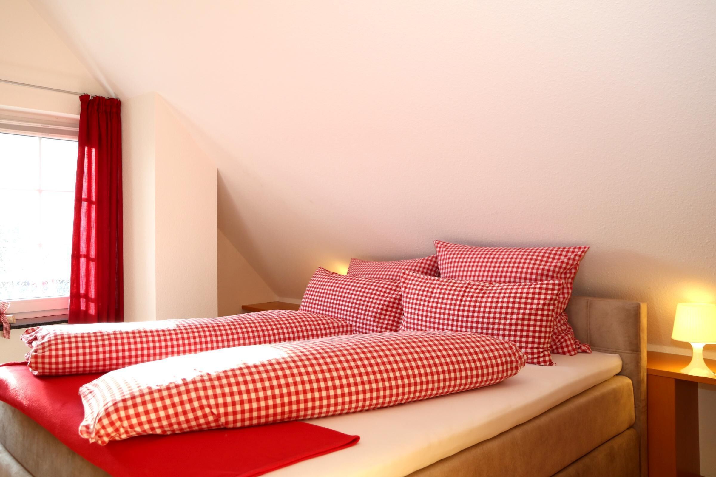 A bedroom with a bed, red checkered bedding, a window with red curtains, and a bedside lamp.
