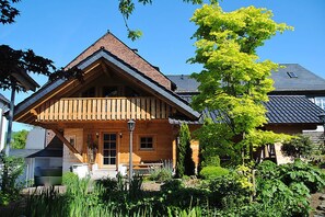 Una casa de madera con techo inclinado, un porche y un jardín con diversas plantas.