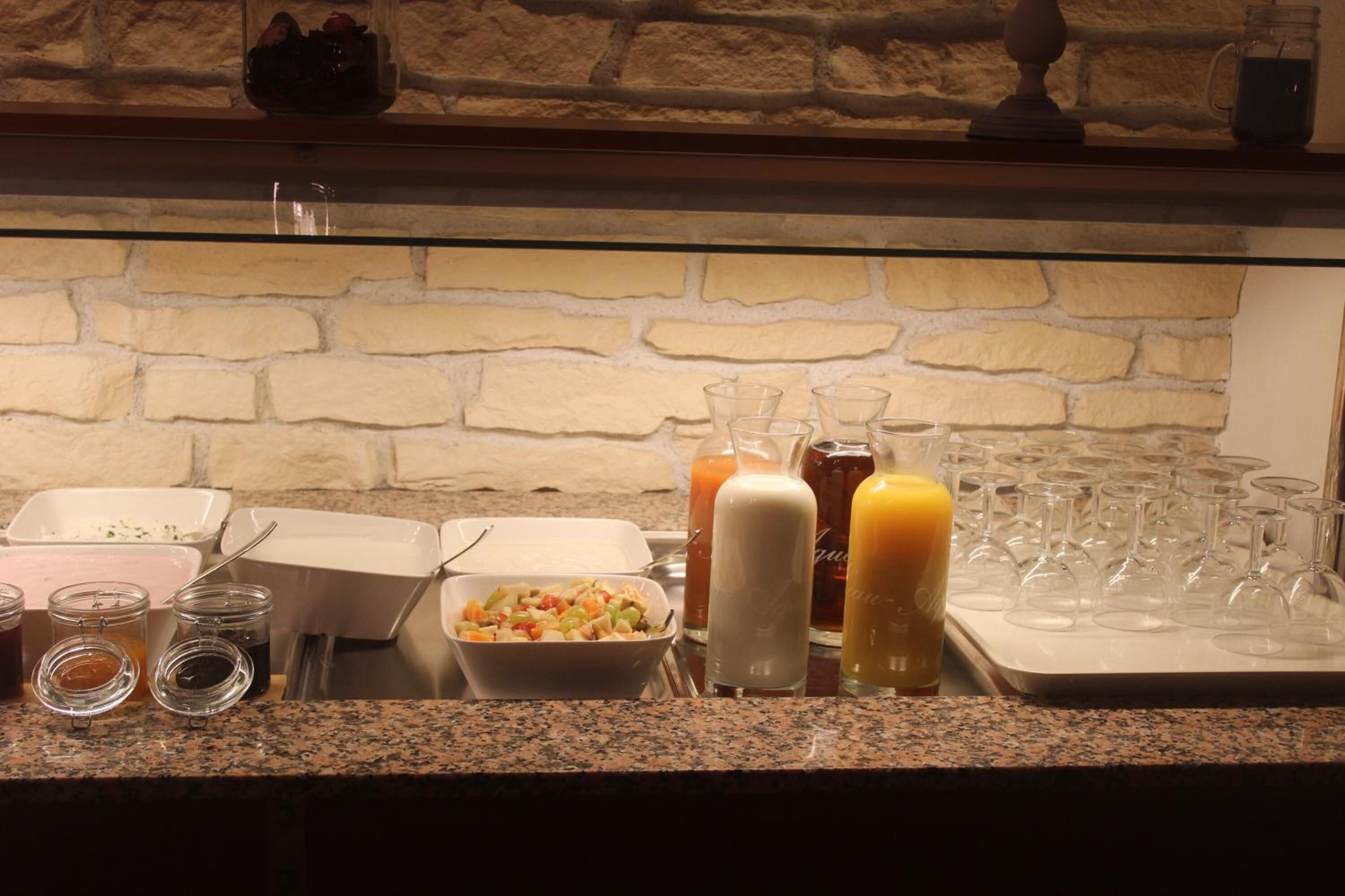 Una barra de desayuno con diversas opciones de comida y bebidas, incluyendo un bol de frutas, condimentos y una bandeja de copas de vino.