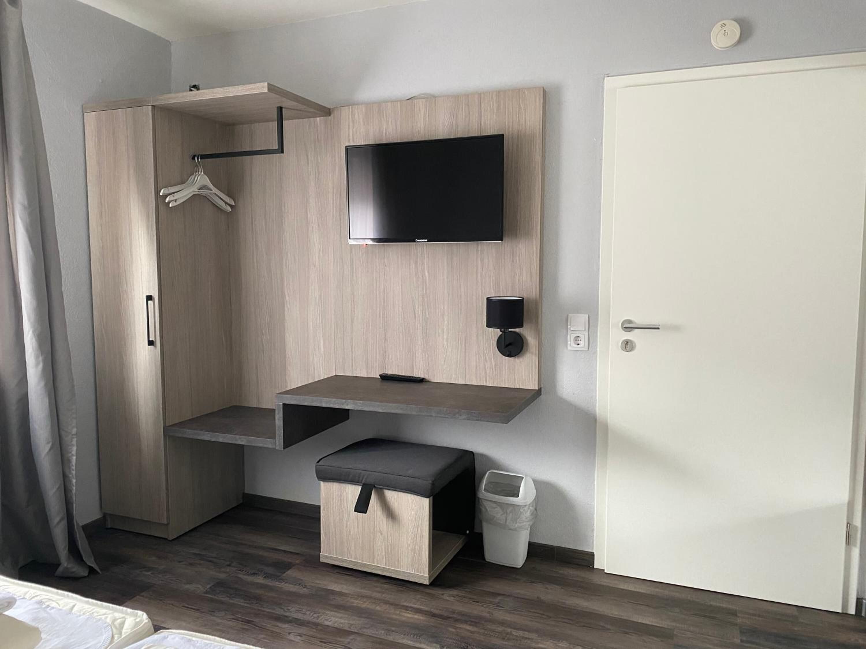 A modern room with a built-in wardrobe, a flat-screen TV mounted on the wall, a small bench, and a waste bin.