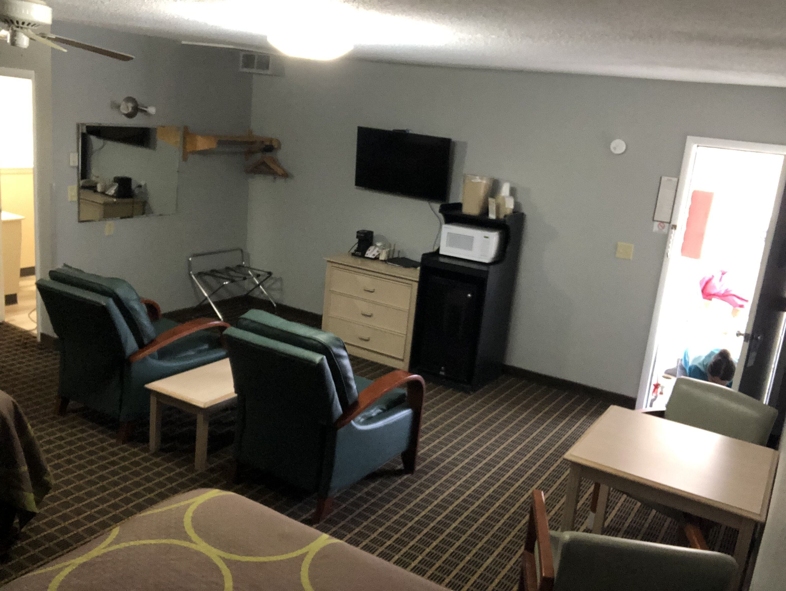 A room with a flat-screen TV mounted on the wall, a small cabinet with a microwave, and a seating area with two chairs and a table.
