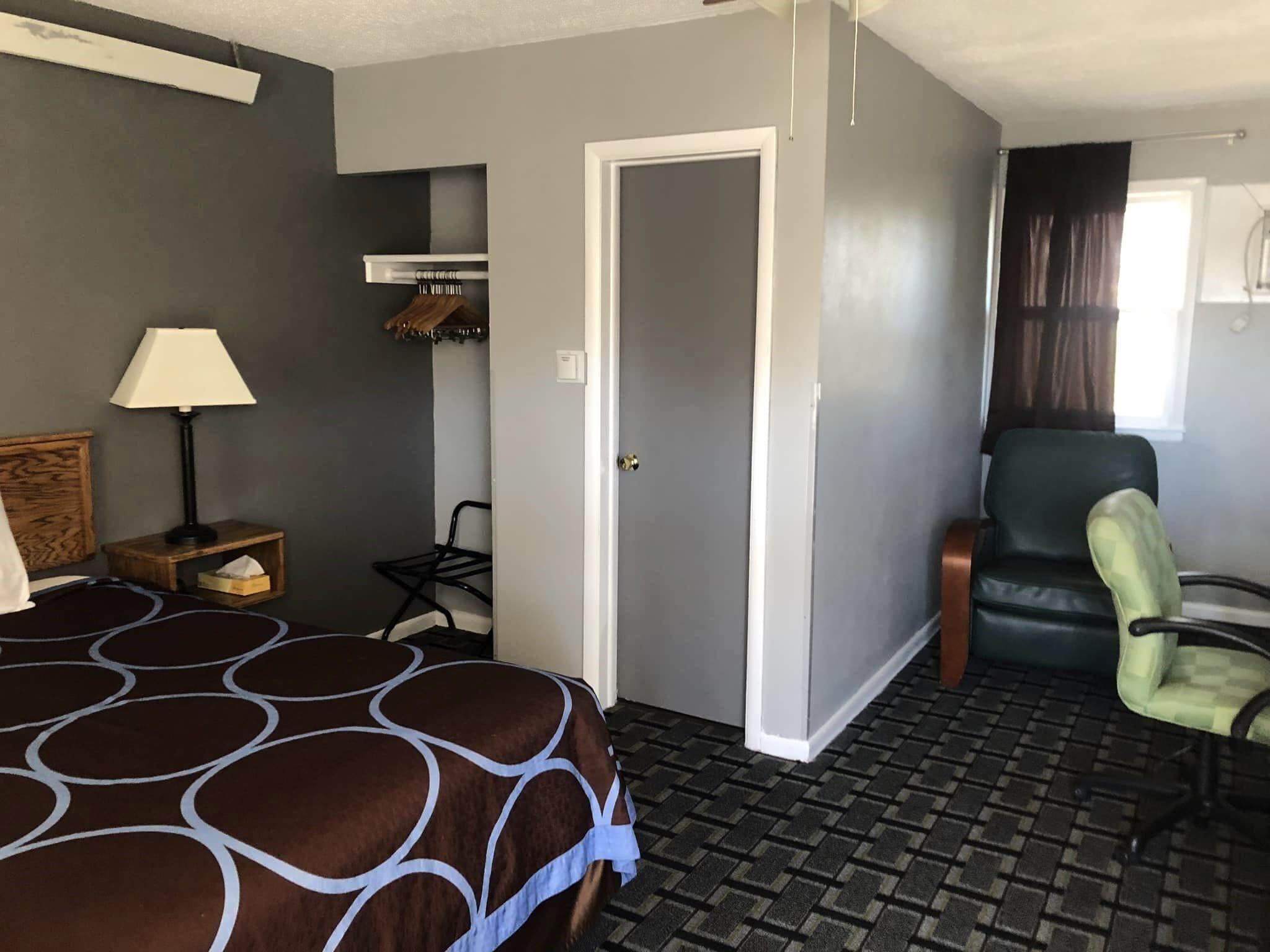 A hotel room with a bed, a chair, a desk with a lamp, a coat rack, and a door.