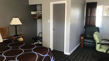 A hotel room with a bed, a chair, a desk with a lamp, a coat rack, and a door.