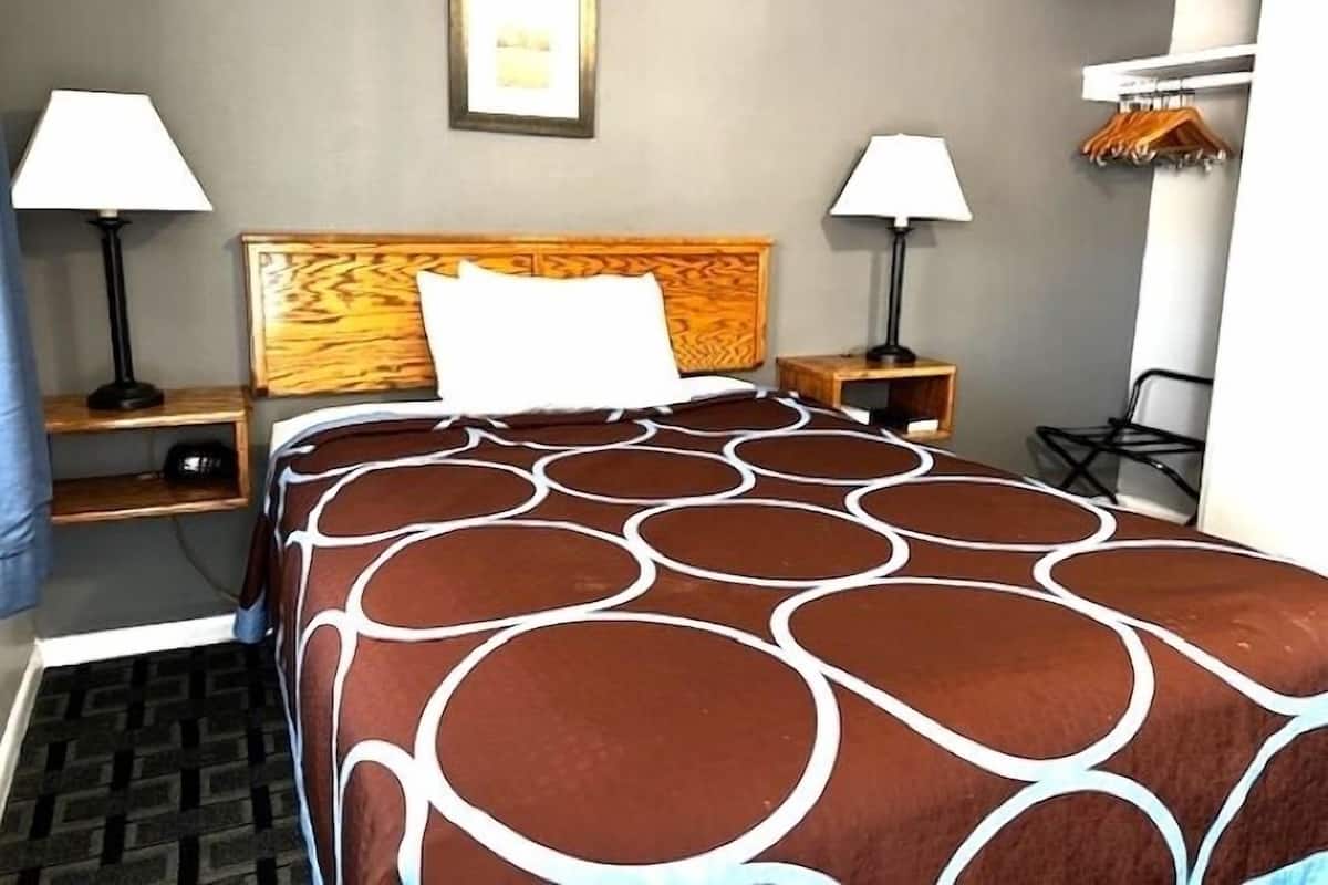 Une chambre d’hôtel avec un lit, deux tables de chevet, des lampes, une tête de lit en bois, une étagère murale avec des vêtements et un tapis à motifs.