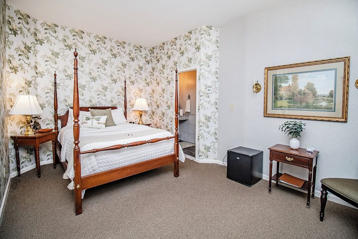 A four-poster bed with a wooden frame, two bedside tables with lamps, a small desk with a chair, a framed picture on the wall, and a carpeted floor.