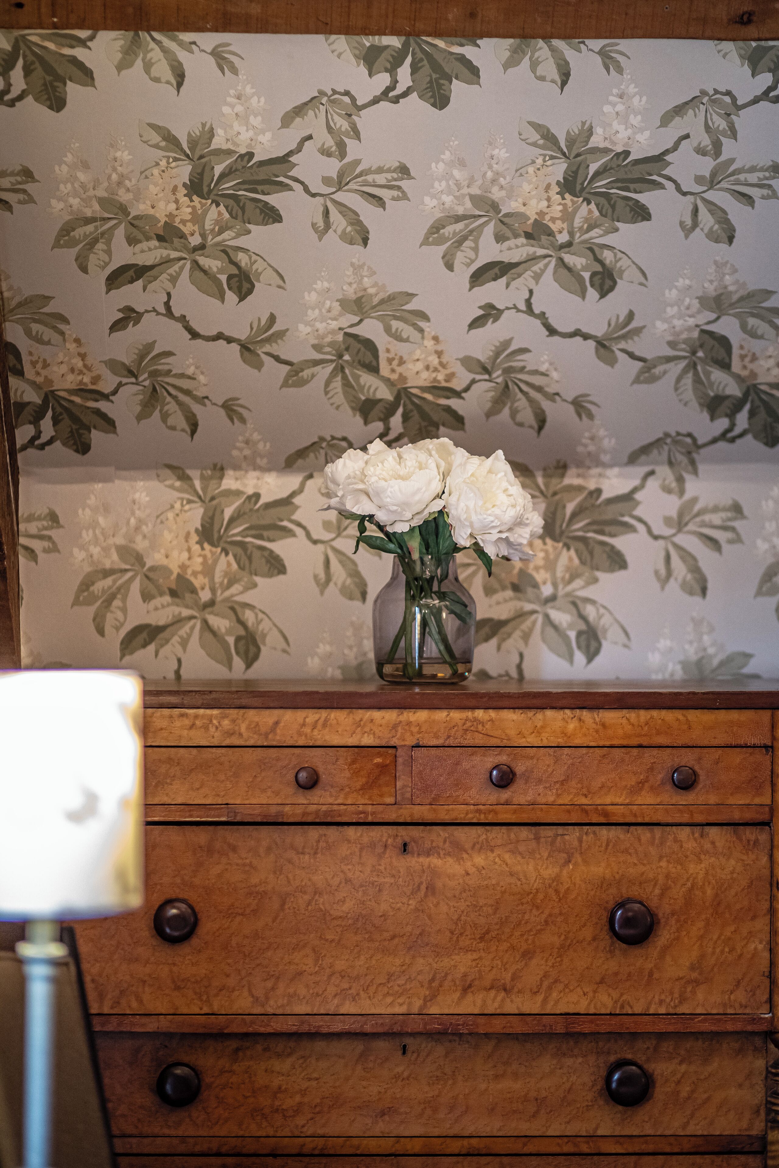 A wooden dresser with a floral wallpaper background and a vase with white flowers on top.