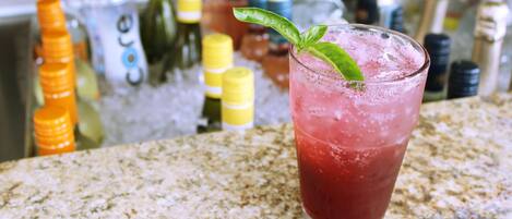 A glass of red beverage with a mint leaf garnish on a granite countertop, surrounded by various bottles and containers.
