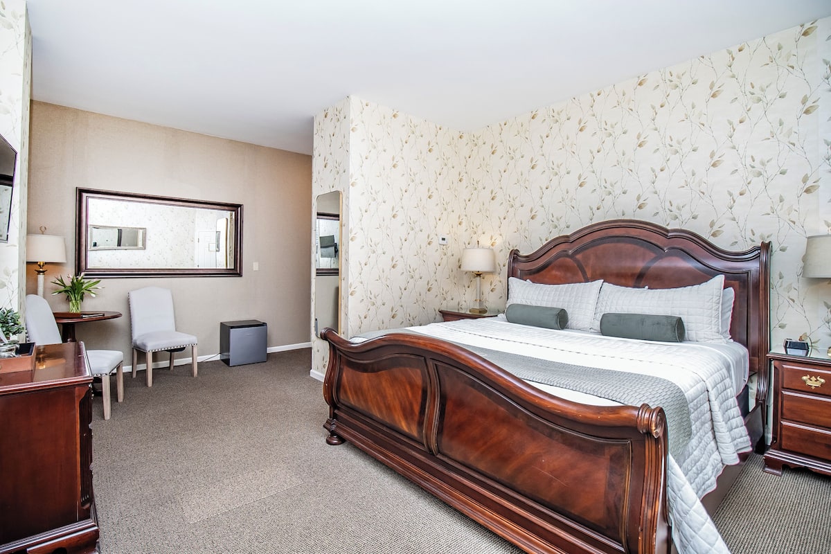 A hotel room with a large wooden bed, a desk, a chair, a small table, and a mirror.