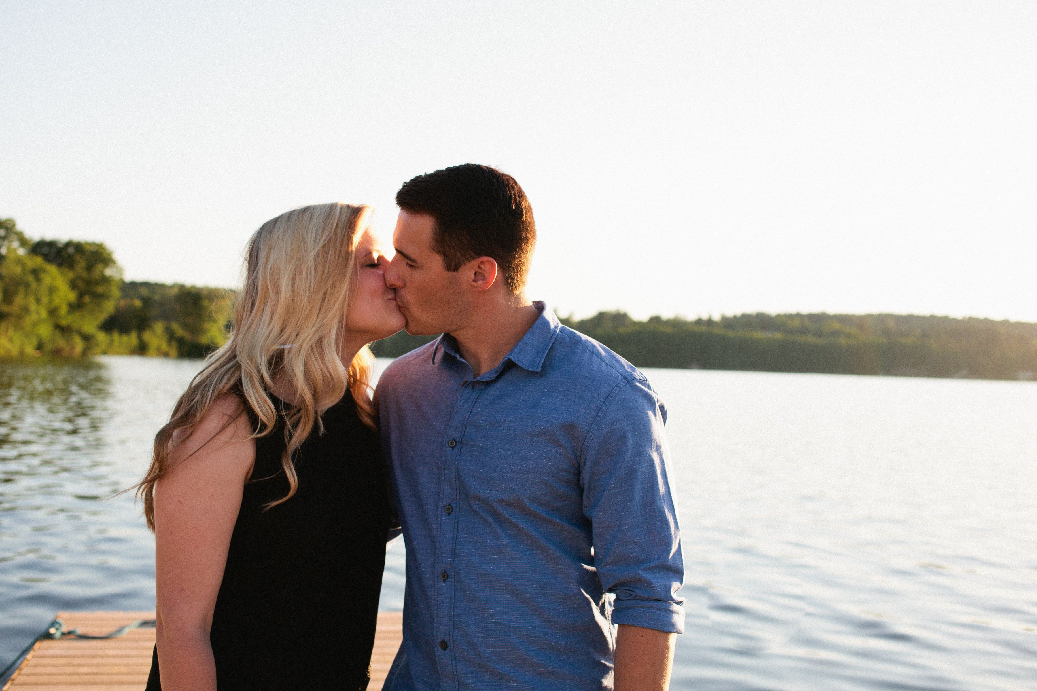 A couple on a wooden dock by a calm lake with trees in the background.