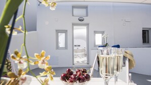 A room with a white interior, a door leading to a balcony, and a table with a glass of champagne and a bowl of cherries. - Ambition Suites (Santorini)