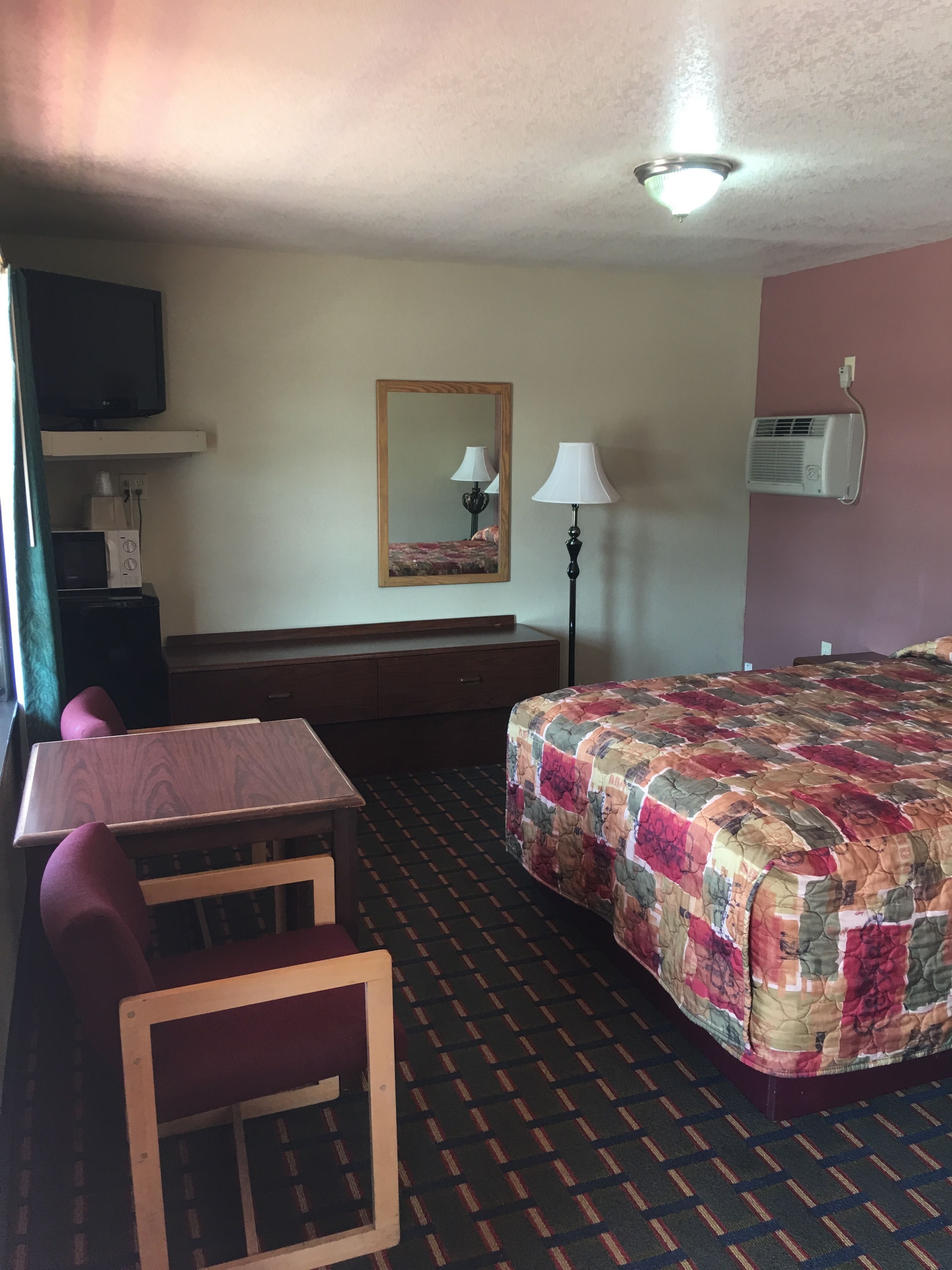 A hotel room with a bed, a desk, a chair, a television, a mirror, and an air conditioning unit.