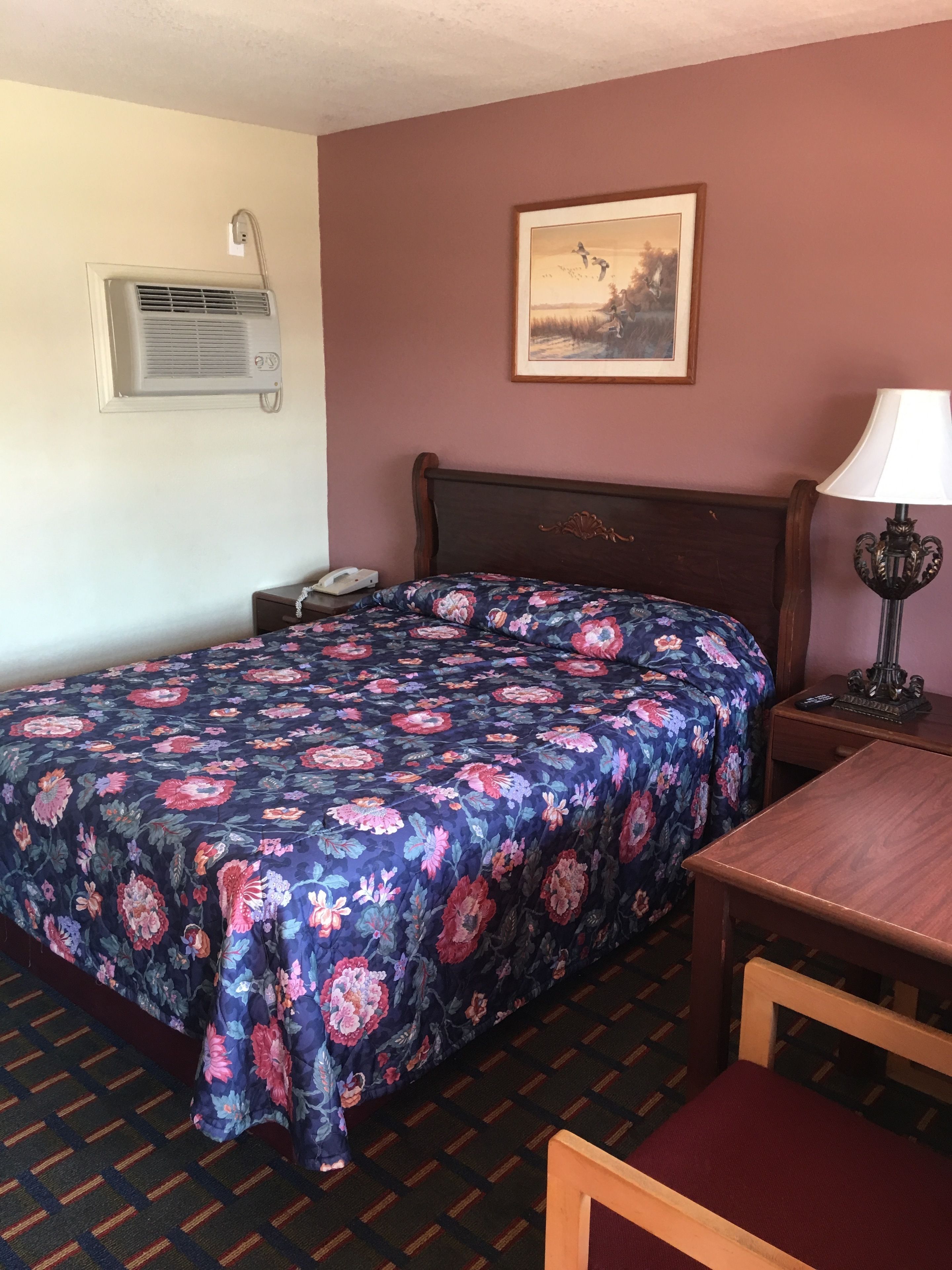 A hotel room with a bed, a nightstand, a lamp, a framed picture on the wall, and an air conditioning unit.