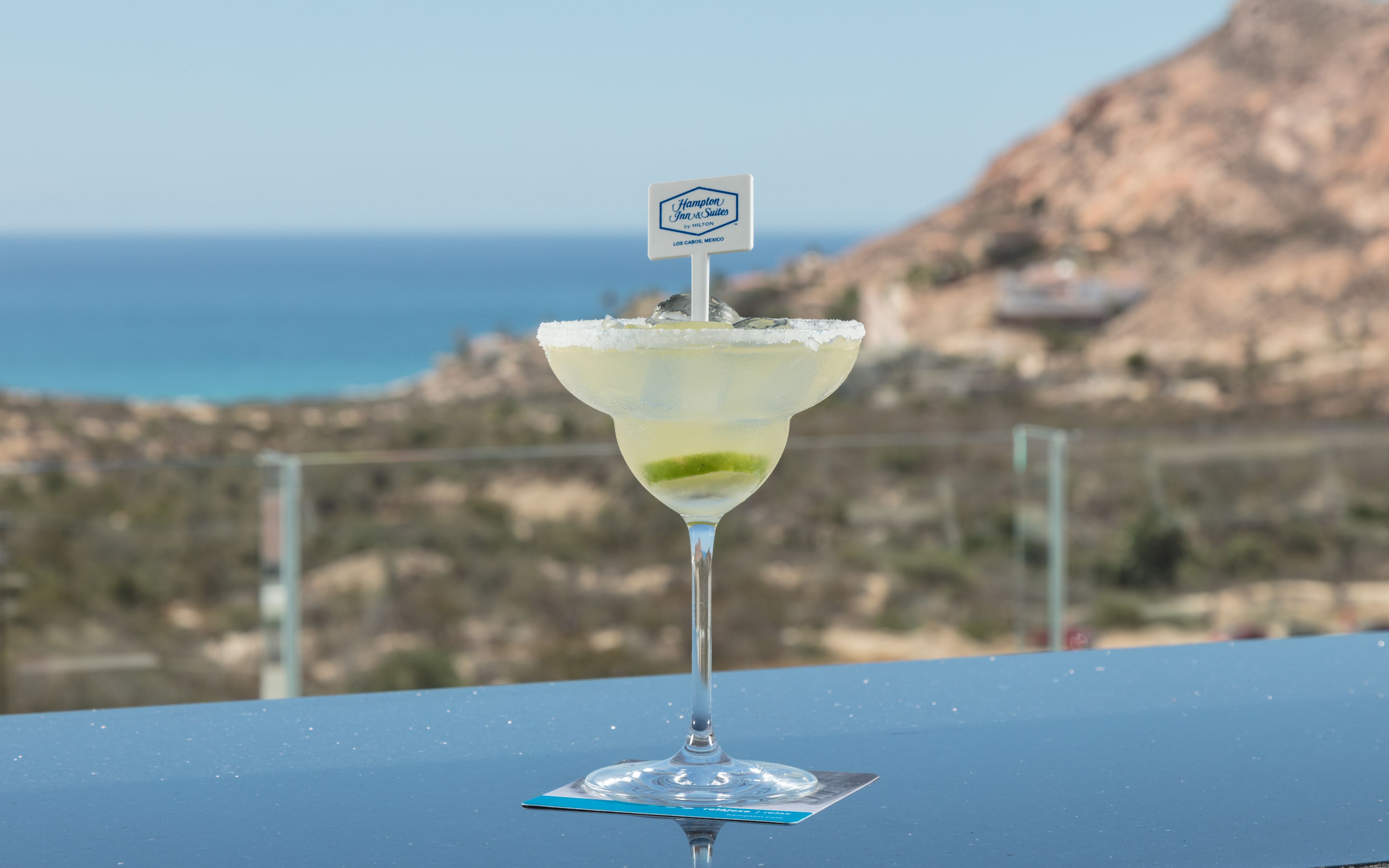 Ein Cocktailglas mit einem Schild auf dem Tisch, vor einer malerischen Aussicht auf das Meer und die Hügel.