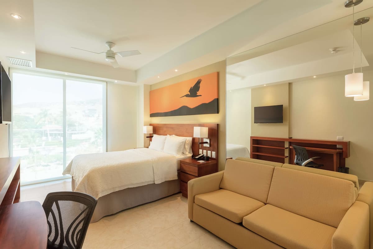 Quarto de hotel com cama, sofá, escrivaninha, cadeira, televisão e um quadro de um pássaro em voo.