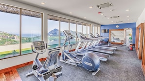 Un gimnasio con cintas de correr, elípticas y un espejo. Cuenta con grandes ventanales que ofrecen vistas al mar y a edificios.