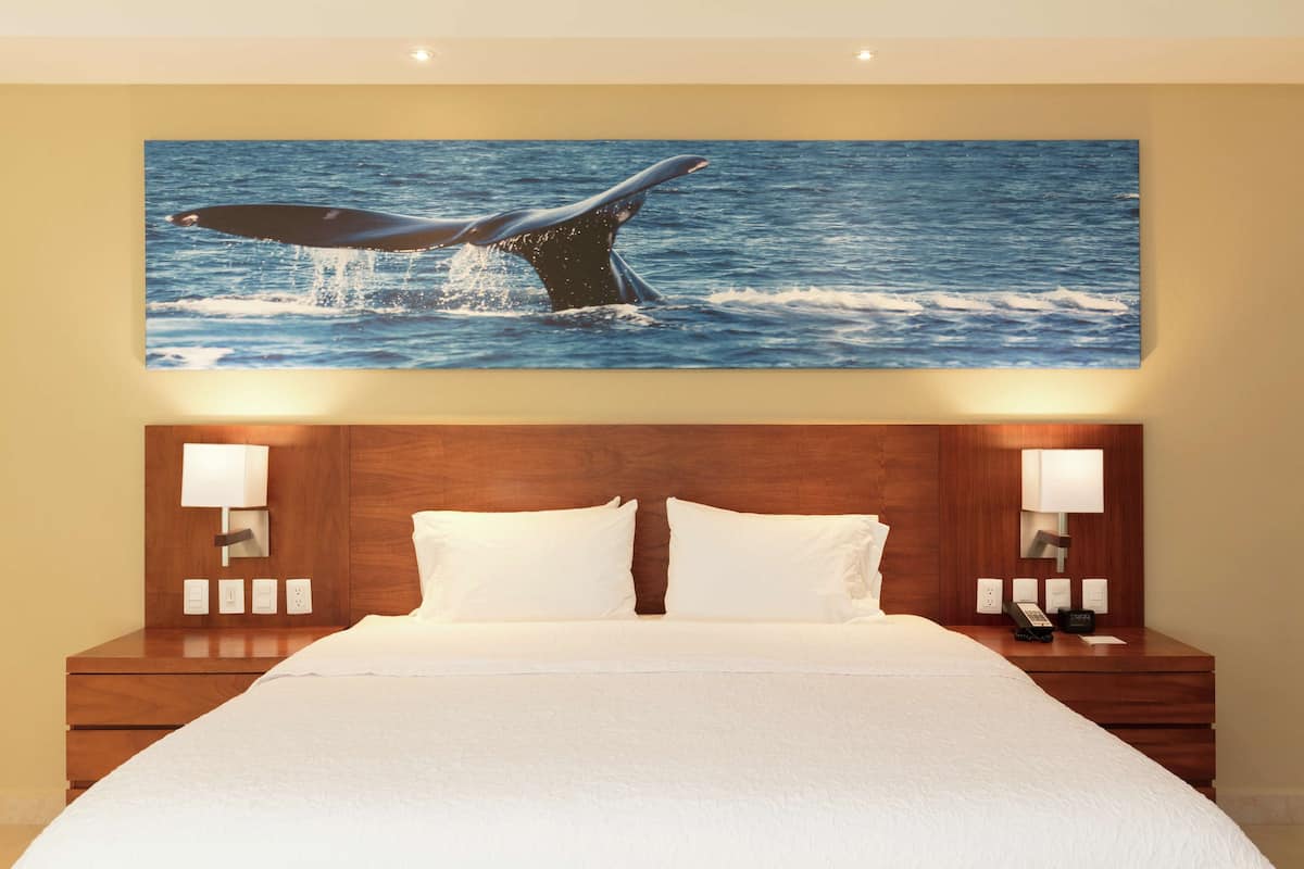 Quarto de hotel com cama grande, cabeceira de madeira, duas luminárias de cabeceira e uma obra de arte na parede com o rabo de uma baleia.