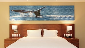Habitación de hotel con una cama grande, cabecera de madera, dos lámparas de noche y una obra de arte en la pared con la forma de una cola de ballena.