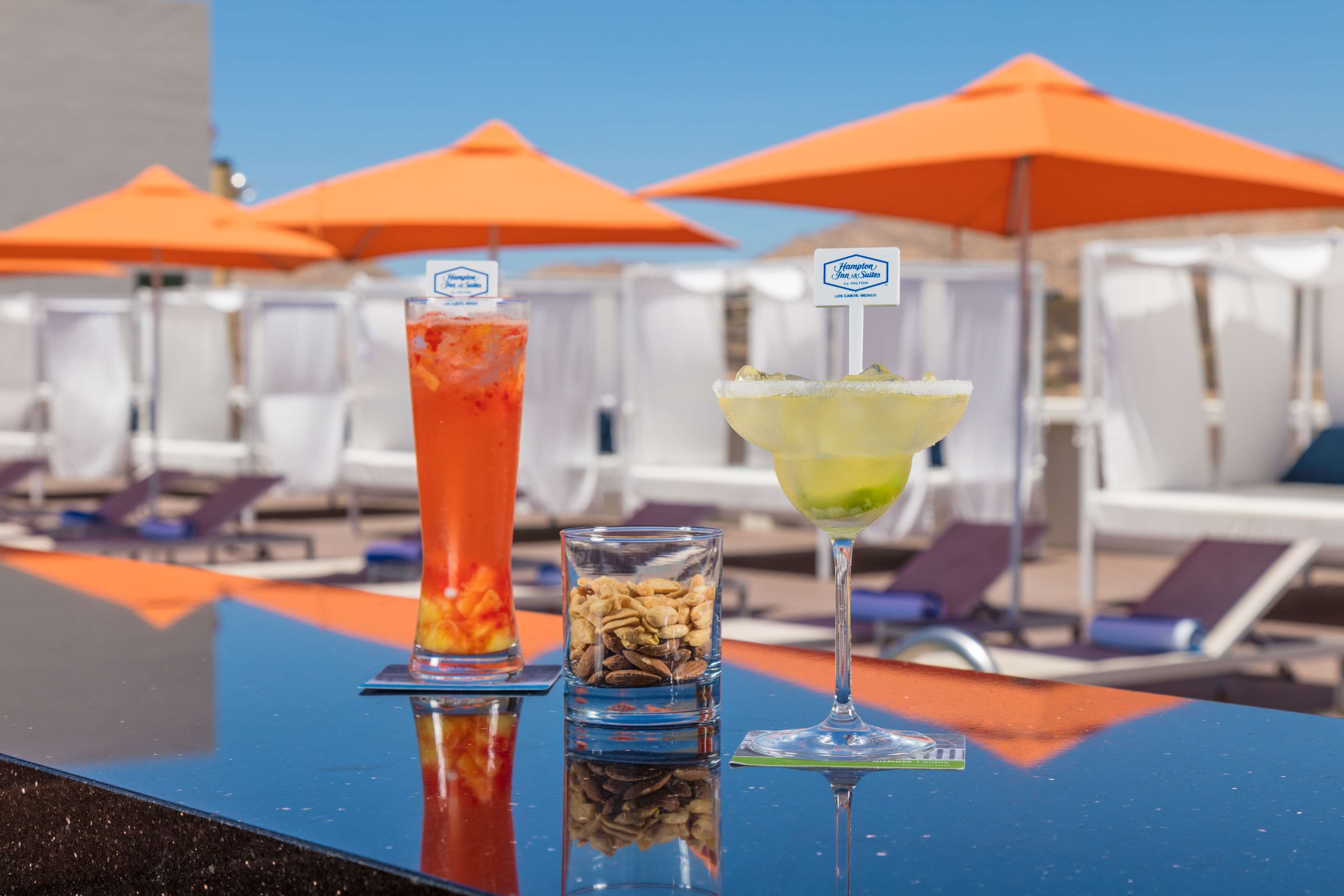 A bar with two cocktails and a glass of nuts, with orange umbrellas and lounge chairs in the background.