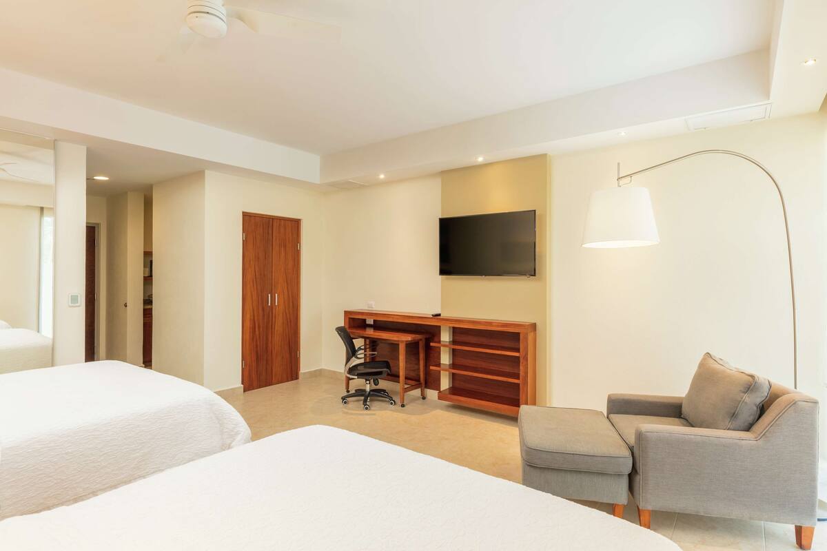 Quarto de hotel com duas camas, uma mesa, uma cadeira, um sofá, uma TV e um abajur.