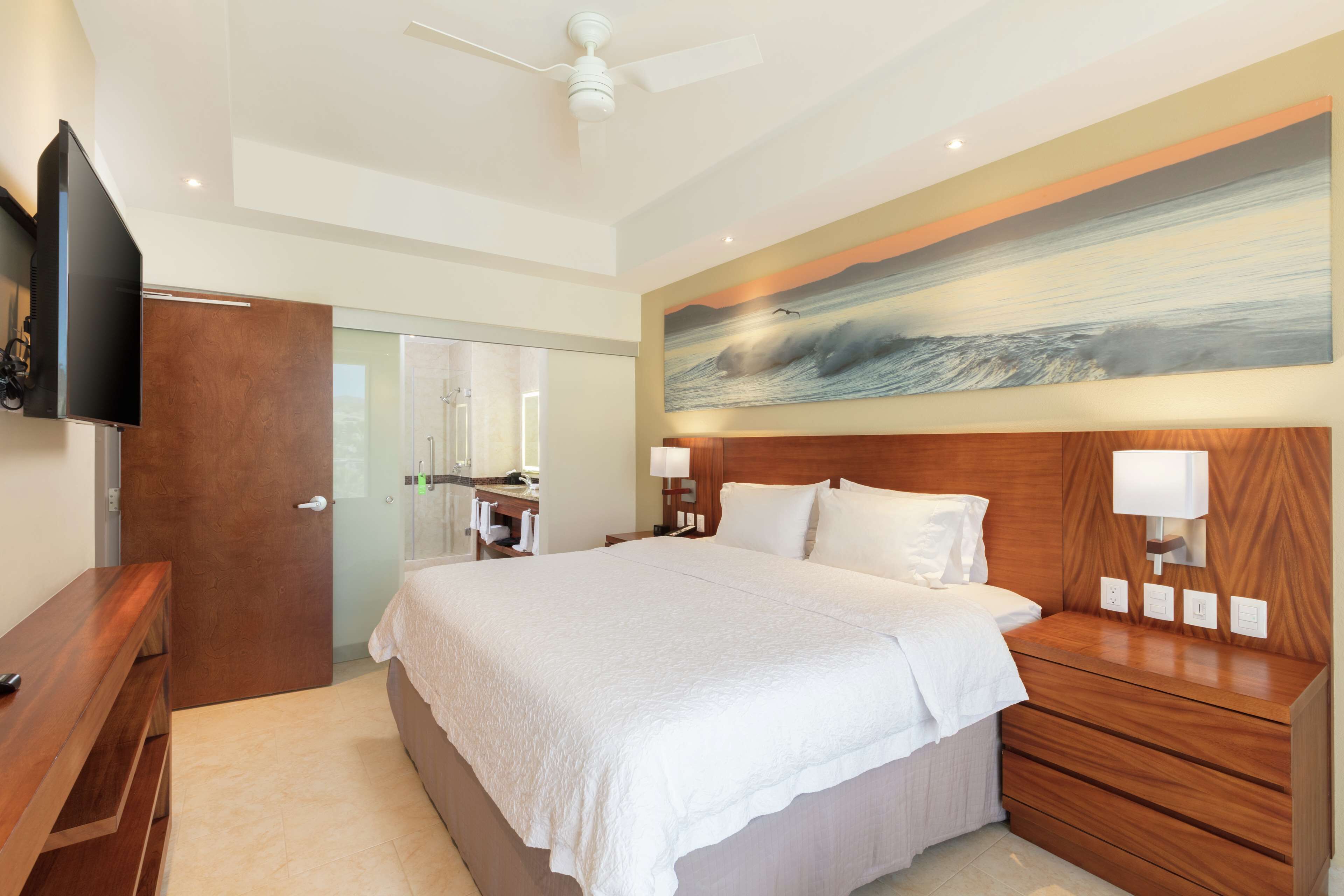 A modern hotel room with a large bed, wooden headboard, bedside lamps, and a painting above the bed.