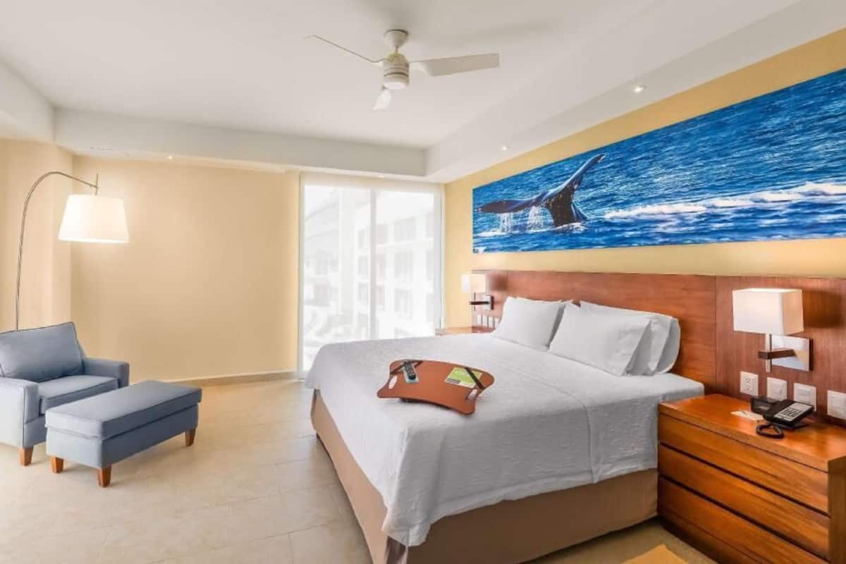 Quarto de hotel com cama, escrivaninha, cadeira, luminária e um grande mural com vista para o oceano.
