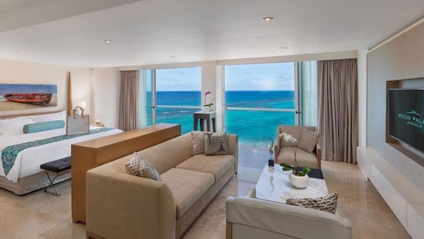 Une chambre d’hôtel avec un lit, un canapé, une table basse et un téléviseur. La chambre dispose de grandes fenêtres donnant sur l’océan.