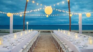 A long table set for a formal event with white tablecloths, candle lanterns, and a view of the ocean.